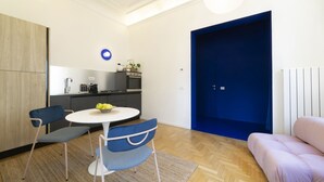 Een moderne woonkamer met een ronde tafel, twee stoelen, een bank, een keukenhoek en een blauwe deur.