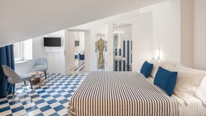 Una habitación de hotel con suelo a cuadros, una cama con ropa de cama a rayas, un televisor montado en la pared y un baño con espejo y azulejos azul rayados.