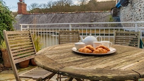 Une table en bois sur laquelle se trouvent une assiette de croissants, une théière et des tasses, le tout placé sur un balcon d’où l’on peut admirer une maison et des arbres.