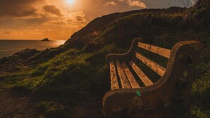 Un banc en bois, placé sur une pente herbeuse, offre une vue sur une zone côtière, avec le soleil se couchant ou se levant à l’horizon.