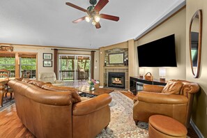 A spacious living room with leather sofas, a fireplace, a flat-screen TV, and a ceiling fan.