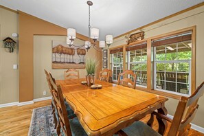 A dining room with a wooden table, chairs, a chandelier, and a large window with a view of greenery.