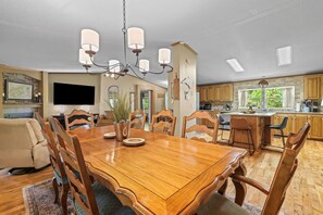 A spacious dining area with a wooden table and chairs, a chandelier, and a kitchen with wooden cabinets and a large window.