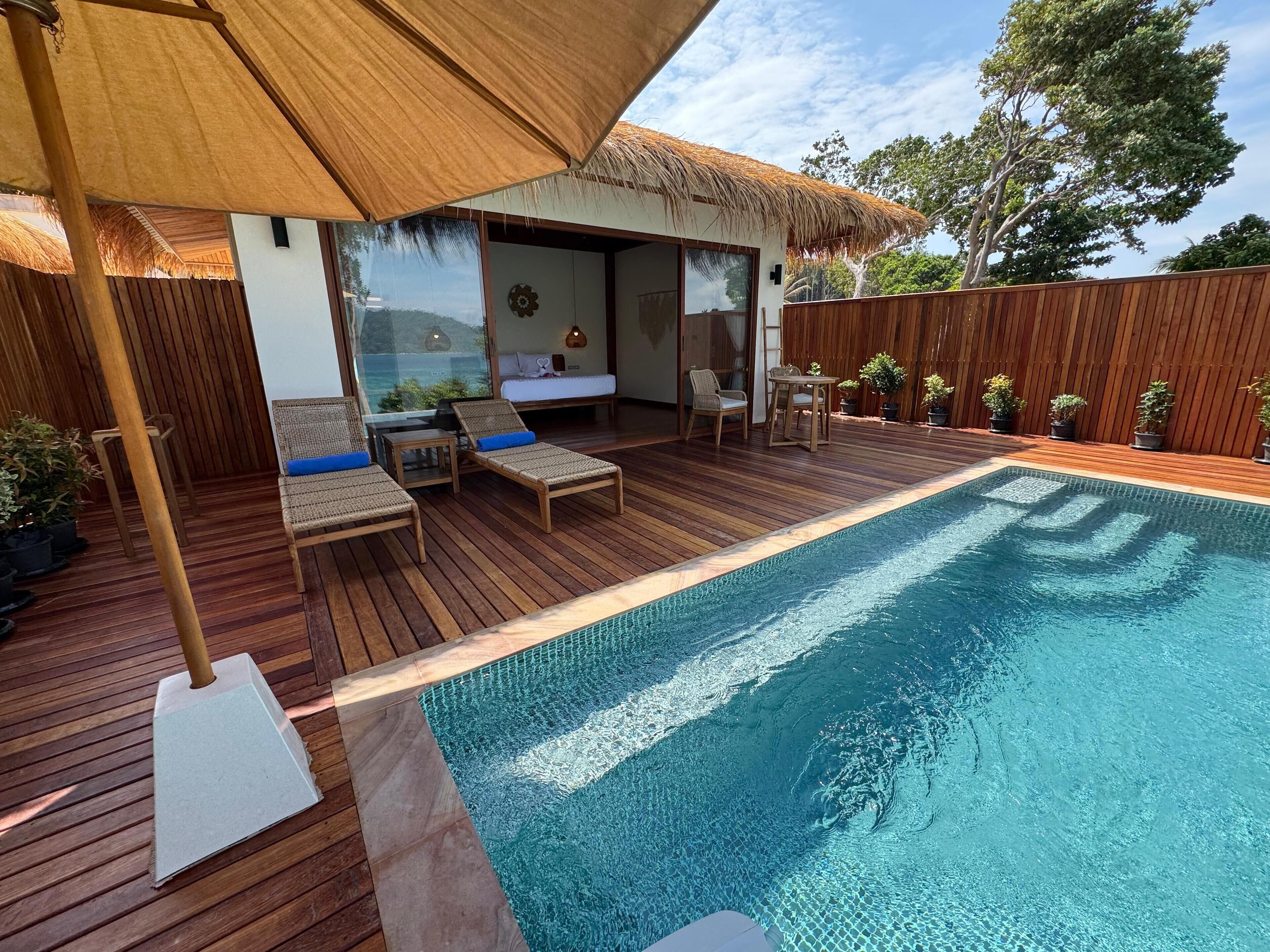 A wooden deck with a pool, lounge chairs, and a thatched roof structure.