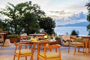An outdoor dining area with wooden tables and chairs, surrounded by greenery and overlooking a body of water. - Ten Moons Sunset Villas (Koh Lipe)