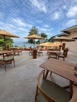 Un patio al aire libre con mesas y sillas de madera, rodeado de sombrillas, con vistas panorámicas al mar y las colinas.