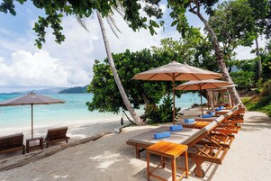 A beachside lounge with wooden tables and chairs, umbrellas, and a view of the ocean.