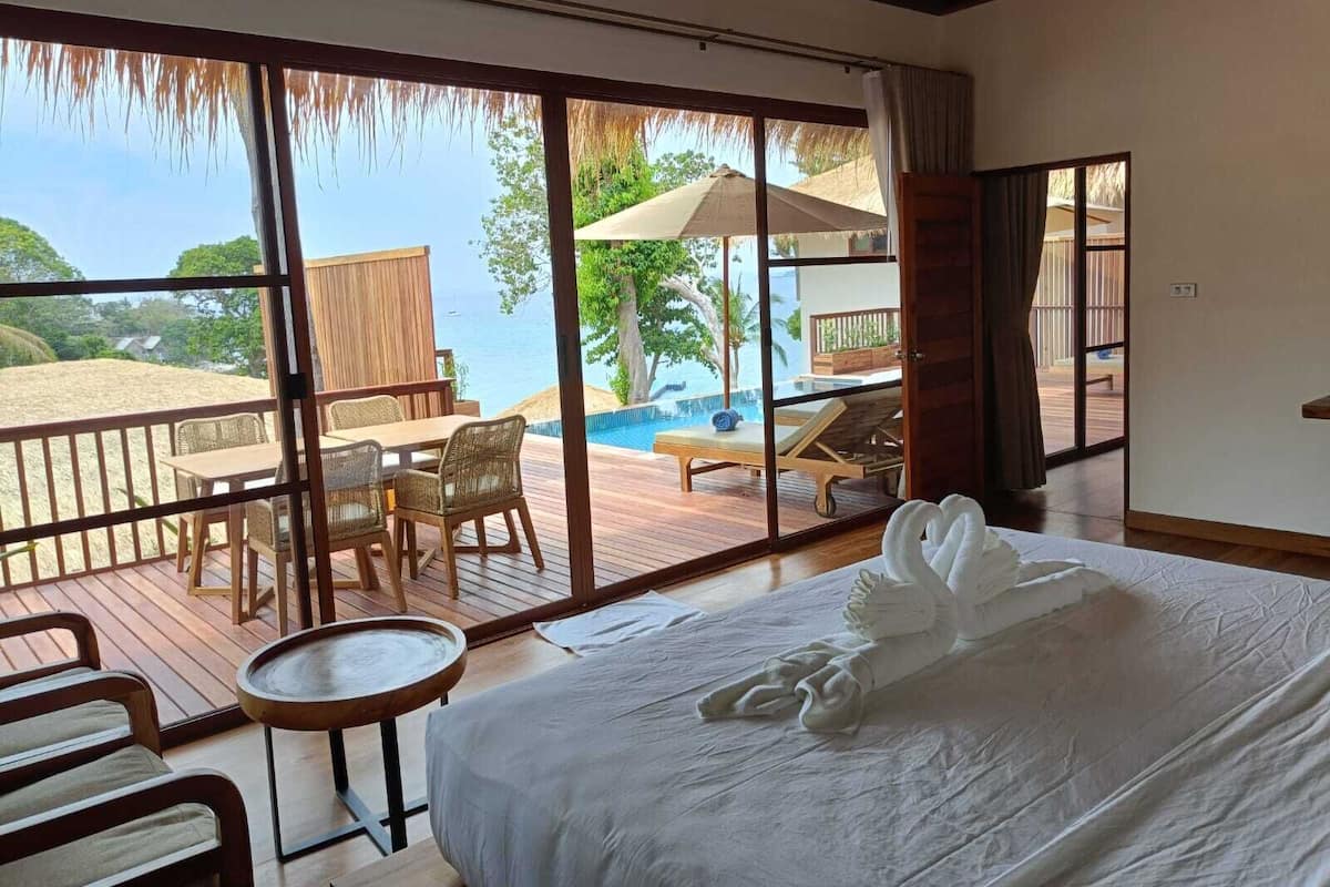 A hotel room with a large bed, a balcony with a view of the beach and pool, and a dining area with a table and chairs.