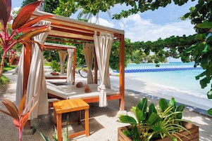 Un salón junto a la playa con toldo de madera, asientos acolchados y vistas al océano.