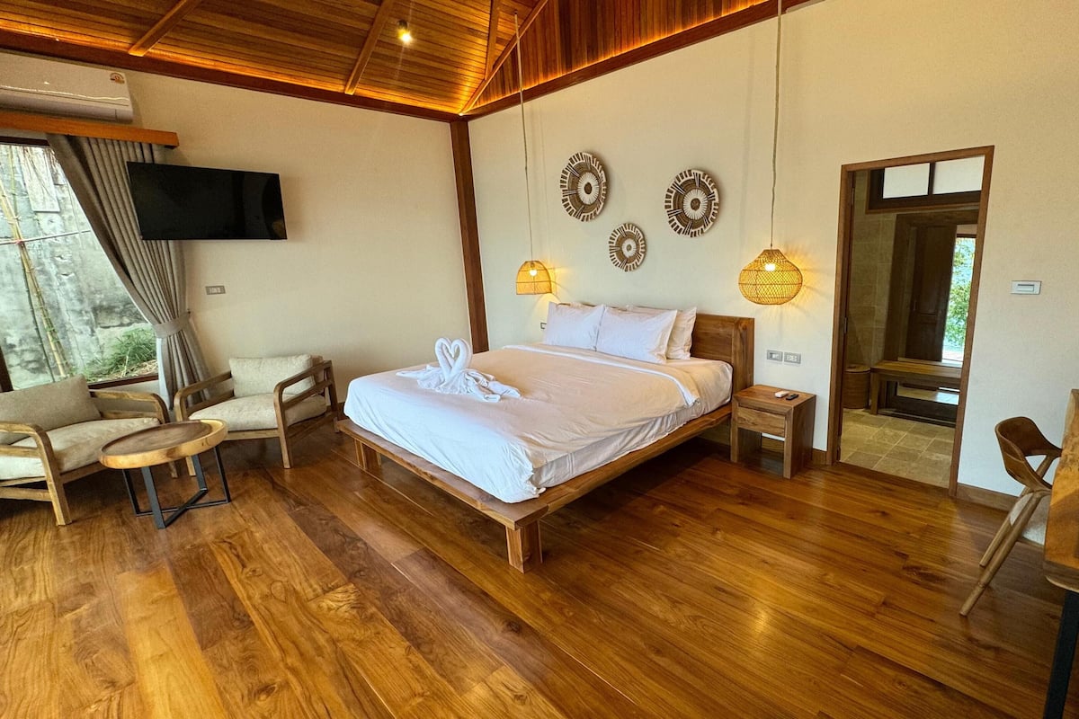 A spacious bedroom with a wooden bed, a sitting area with chairs, a TV mounted on the wall, and a mirror reflecting the room.