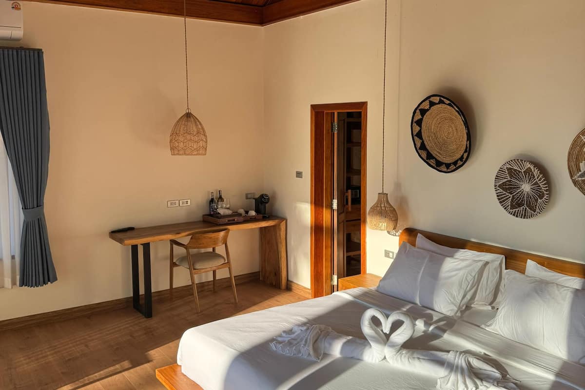 A cozy bedroom with a wooden ceiling, a bed with white linens, a desk with a chair, and a small shelf with decorative items.