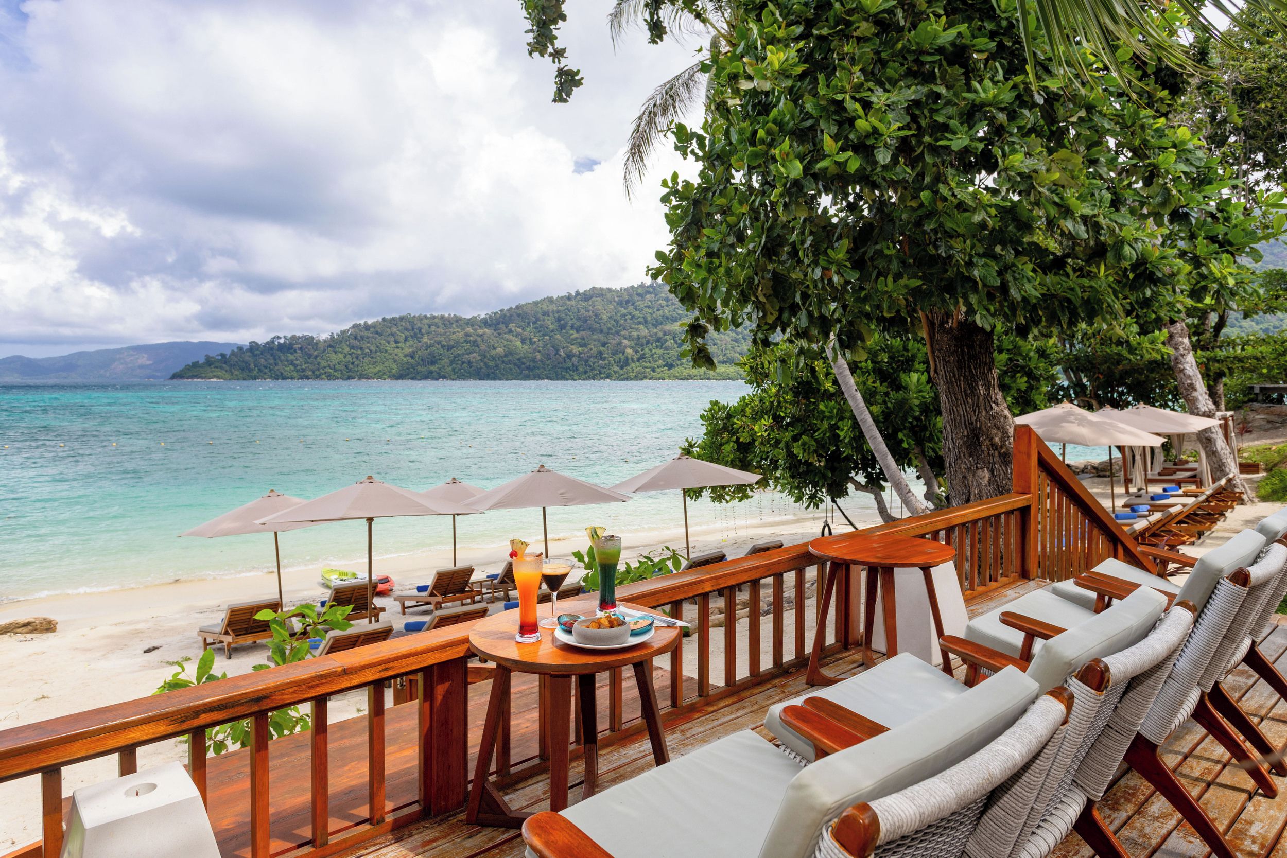 Eine Strandlounge mit Holzmöbeln, Sonnenschirmen und Blick auf das Meer sowie eine bewaldete Insel.
