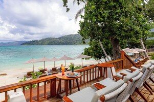 A beachside lounge with wooden furniture, umbrellas, and a view of the sea and a forested island.