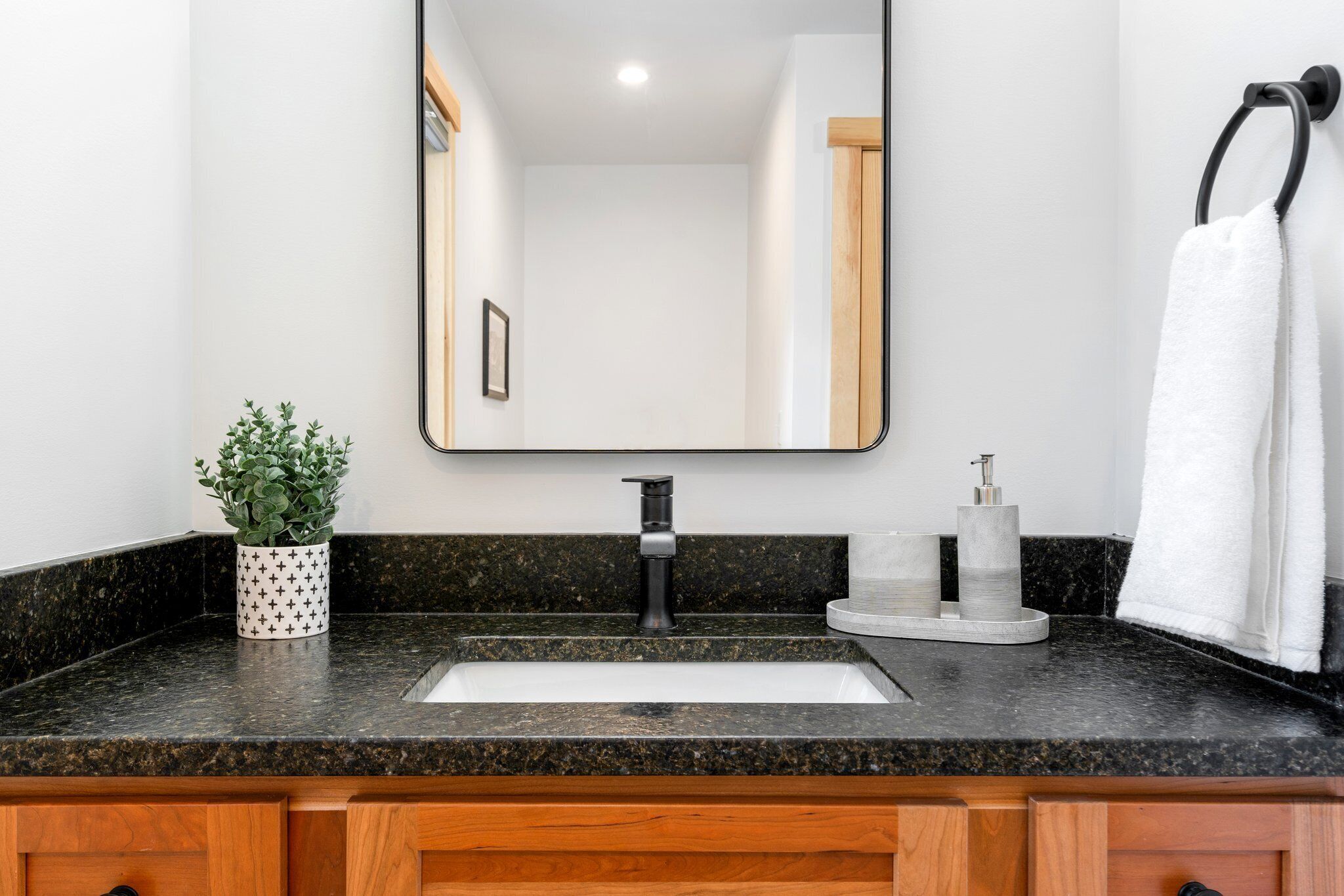 A bathroom with a granite countertop, a white sink, a black faucet, a mirror, a towel rack, and a potted plant.