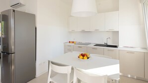 Une cuisine moderne avec des meubles blancs, un réfrigérateur en acier inoxydable, une table à manger blanche avec deux chaises et un panier de fruits posé sur la table.