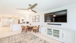 A modern kitchen with white cabinetry, a dining area with wicker chairs, a large TV mounted on the wall, and a ceiling fan.