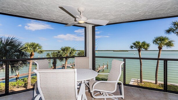 A balcony with a table and chairs, overlooking a body of water with palm trees and a distant shoreline.