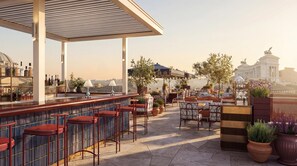 A rooftop bar with outdoor seating, a bar counter, and potted plants.
