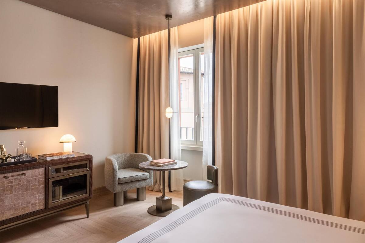 A hotel room with a bed, a small table with a lamp, a chair, a TV mounted on the wall, and a window with curtains.