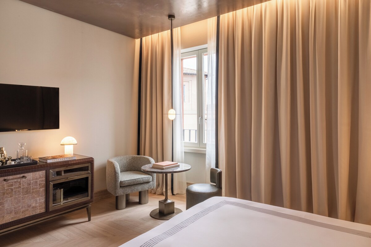 A hotel room with a bed, a small table with a lamp, a chair, a TV mounted on the wall, and a window with curtains.