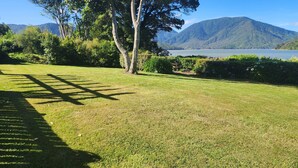 A well-maintained grassy lawn with a wooden pergola casting shadows, surrounded by lush greenery and a backdrop of mountains and a lake. - Waterfront & spacious. Room for cars & boat trailer. 6KM from Havelock Marina (Havelock)