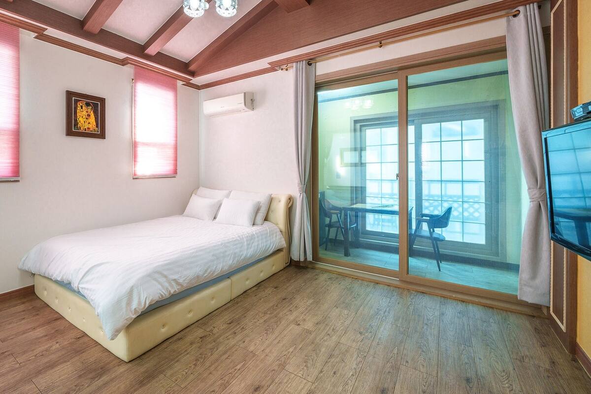 A bedroom with a bed, a sliding glass door leading to a balcony, a framed picture on the wall, and a ceiling with wooden beams.