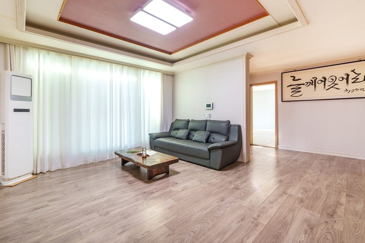 A spacious living room with a grey sofa, a wooden coffee table, and a large window with sheer curtains.
