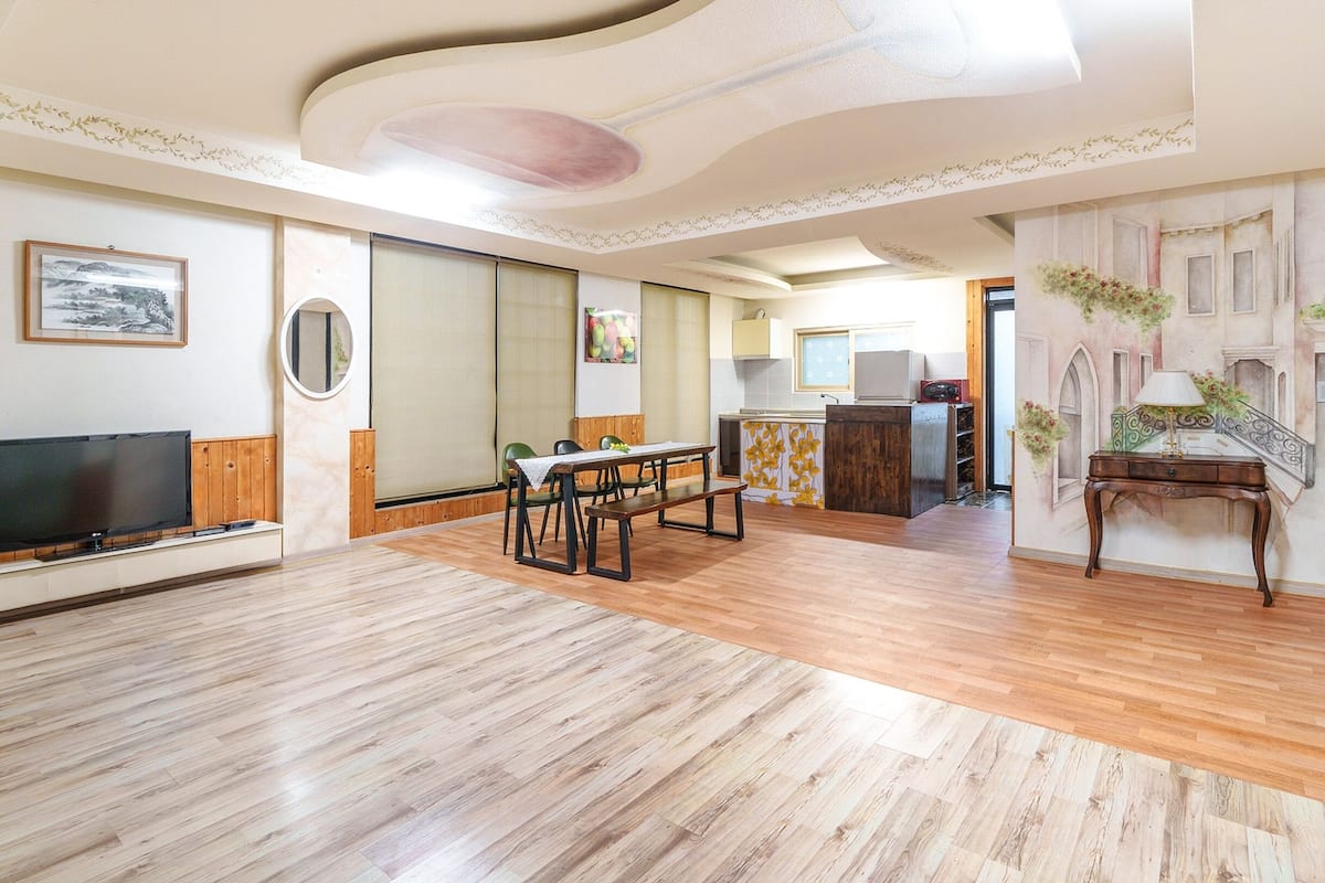 A spacious living area with a wooden floor, a dining table, a TV, and a kitchen in the background.
