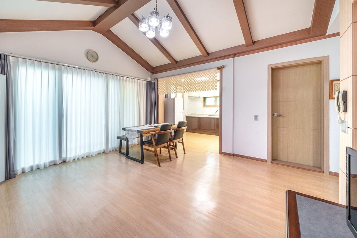 A spacious living area with a dining table, chairs, a kitchen in the background, and a wooden floor.