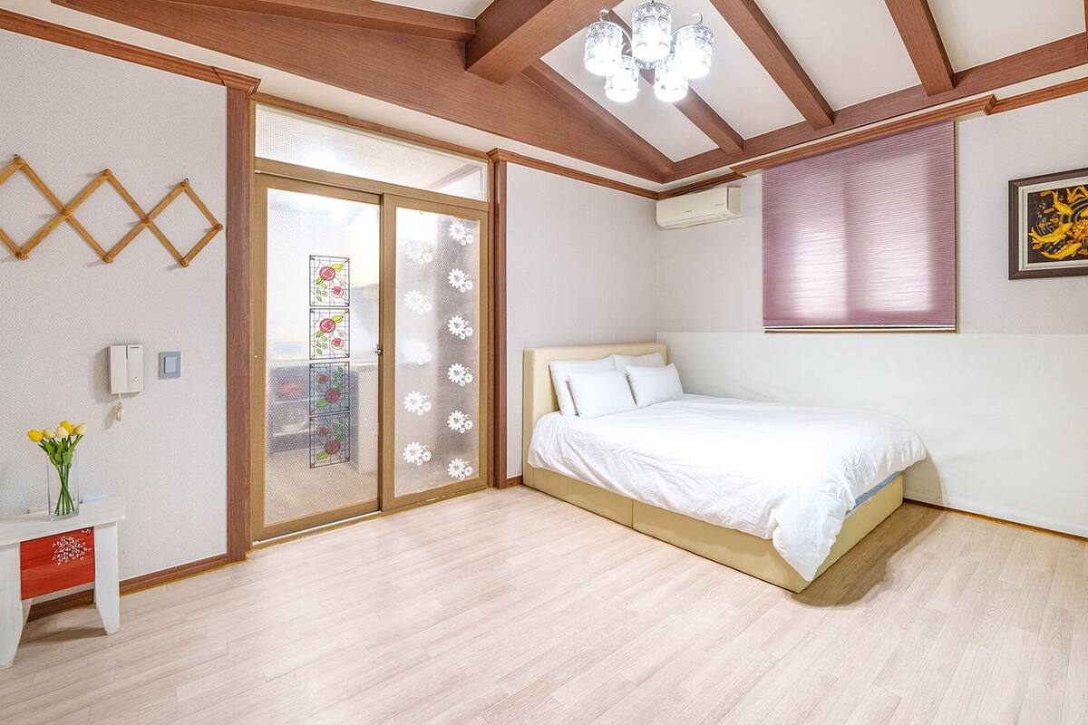 A bedroom with a bed, wooden ceiling beams, a window with blinds, a wall-mounted air conditioner, and a framed picture.