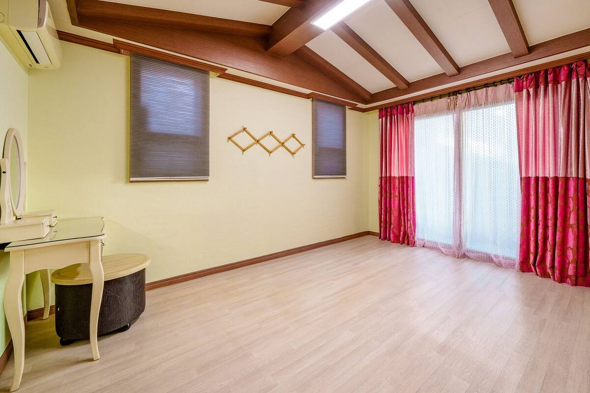 A room with wooden flooring, a desk with a chair, two windows with blinds, and a large window with pink curtains.