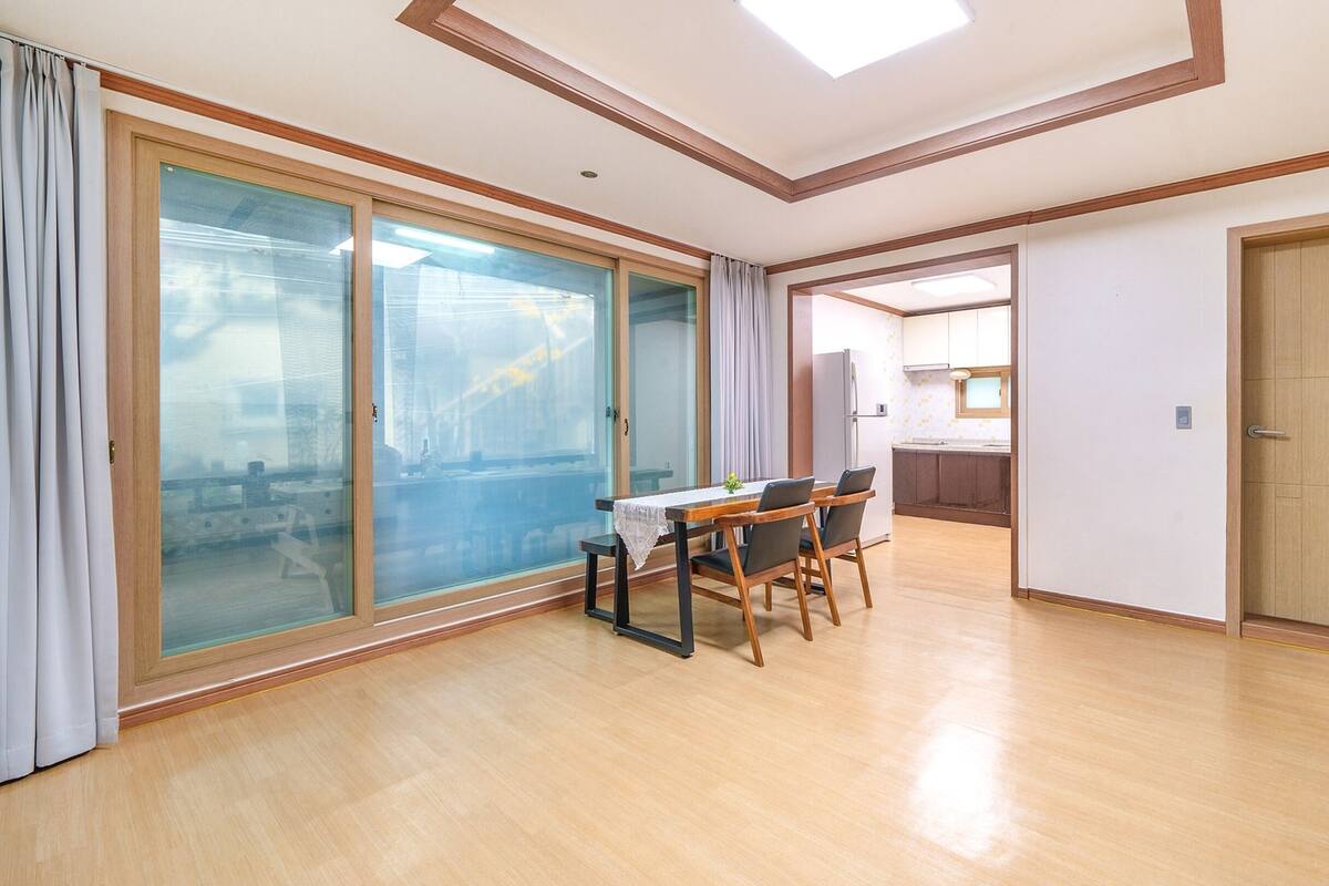 A spacious room with a wooden floor, a dining table with chairs, a sliding glass door, and a kitchen area visible through an open doorway.