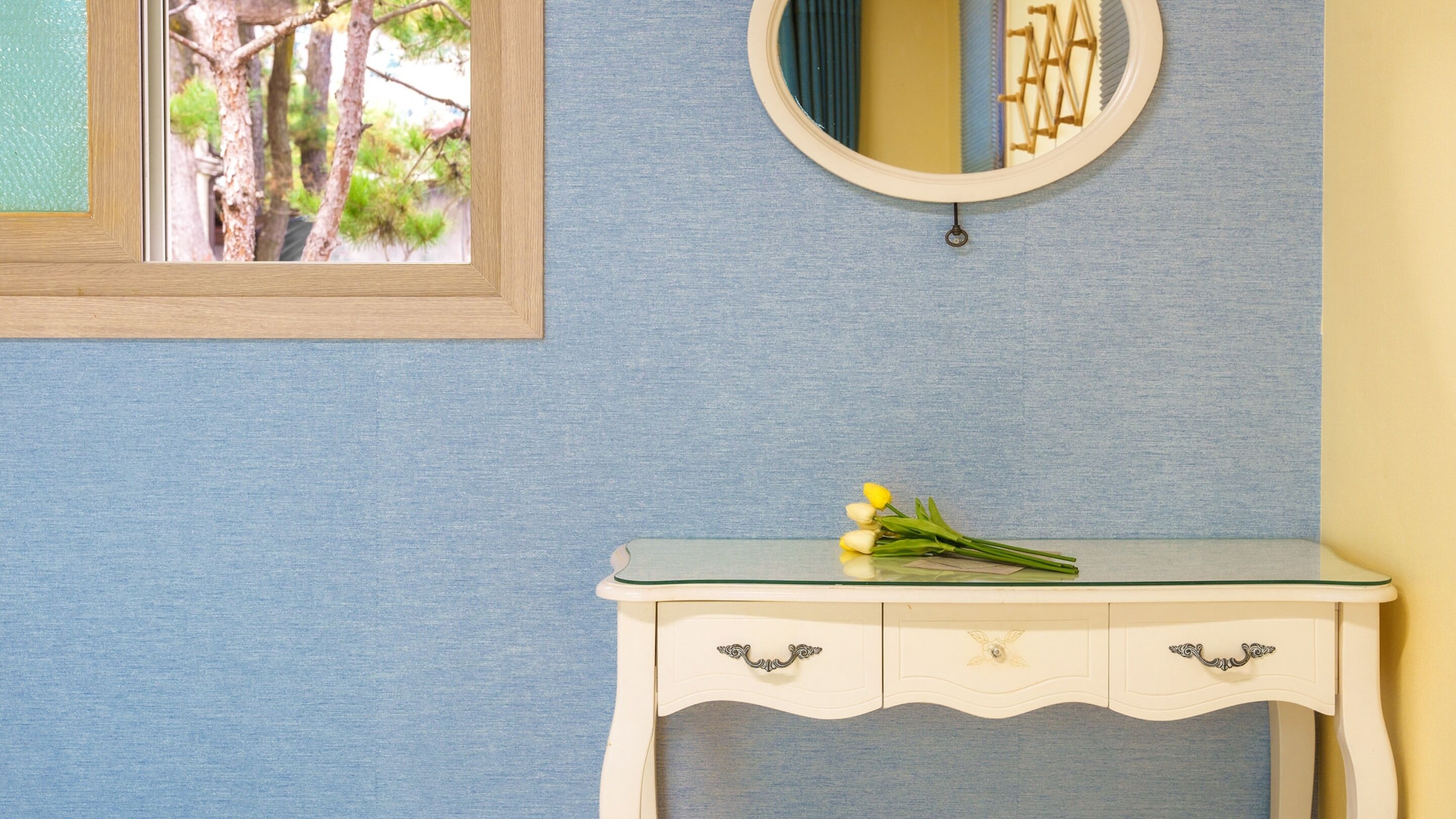 Une pièce avec un mur bleu, une fenêtre à cadre en bois, une commode blanche avec un miroir et un bouquet de fleurs jaunes posé sur la commode.
