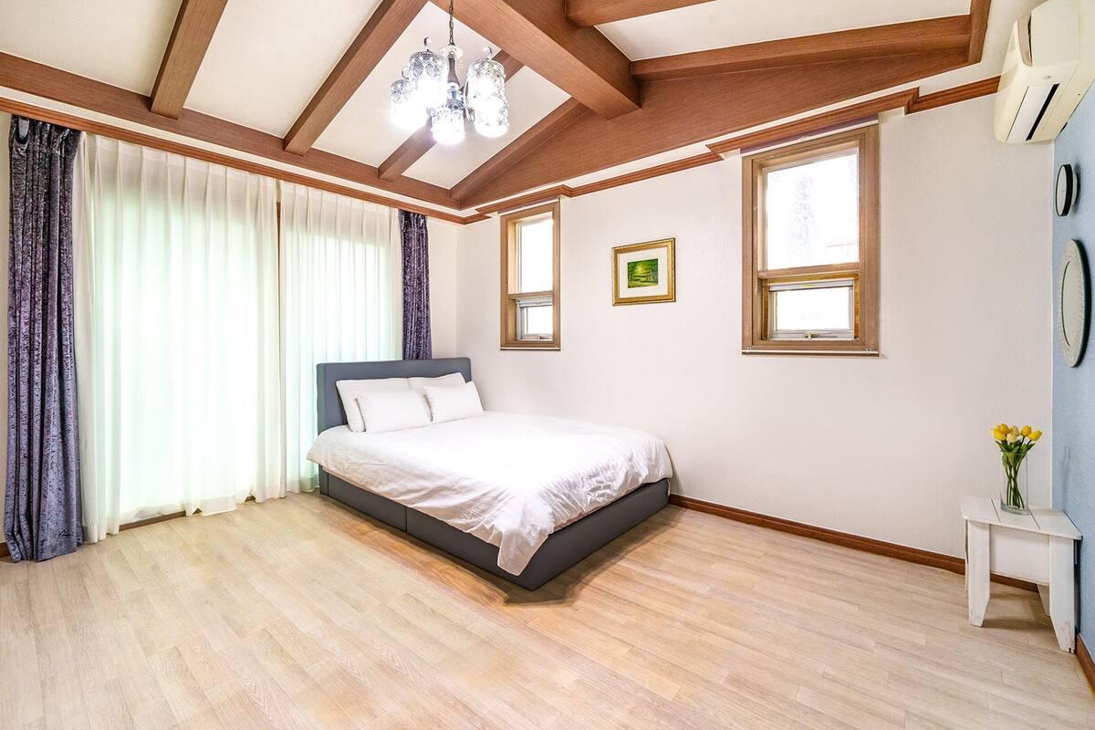 A bedroom with a bed, wooden beams, a chandelier, a window, and a small table with flowers.