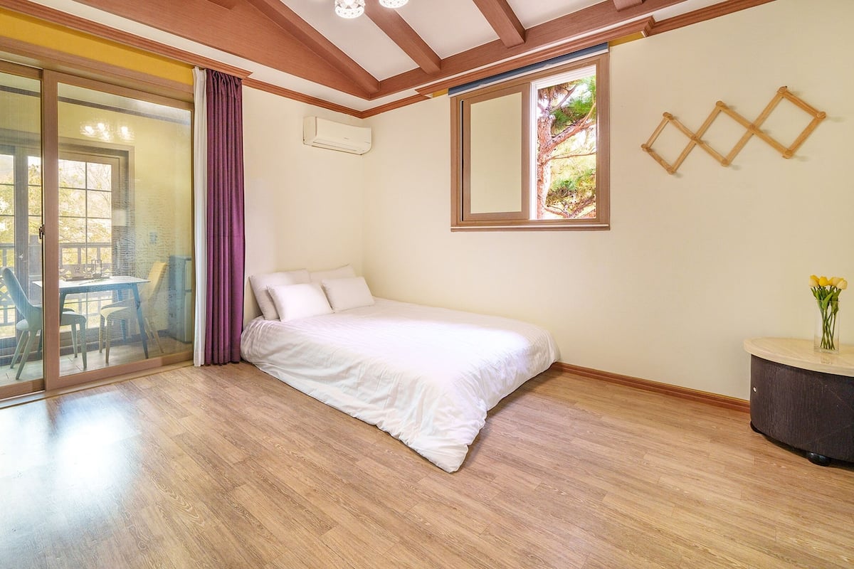 A bedroom with a bed, a sliding glass door leading to a balcony, a small table with a vase of flowers, and a window with a view of trees.