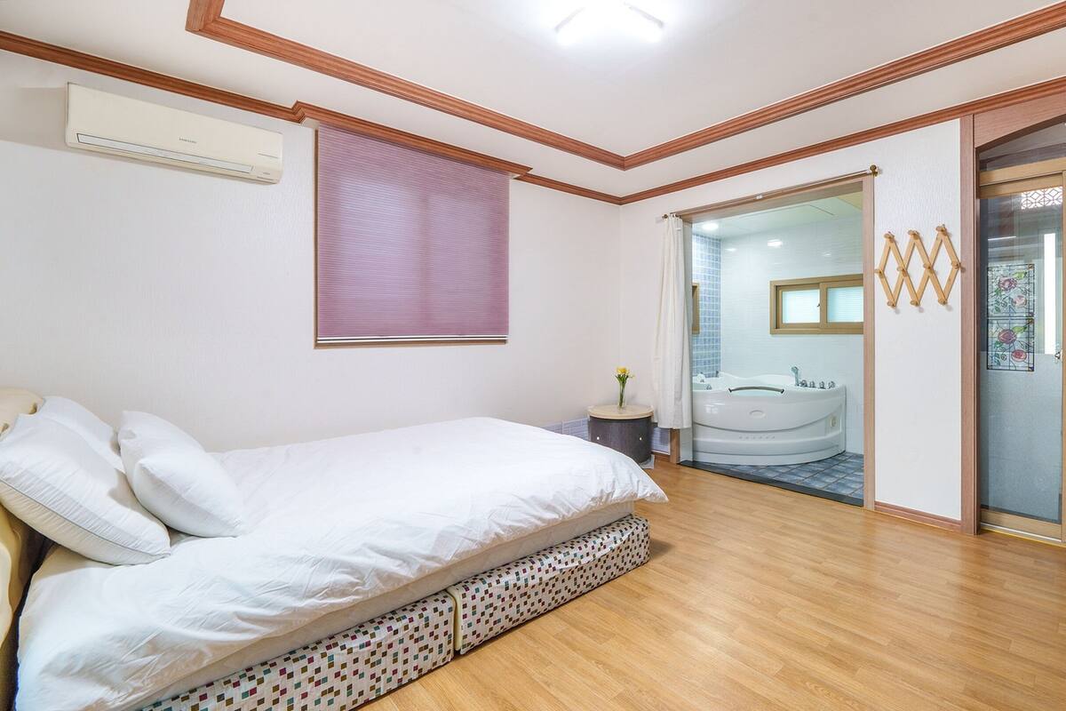 A bedroom with a bed, a window with blinds, a bathroom with a bathtub, and a TV.