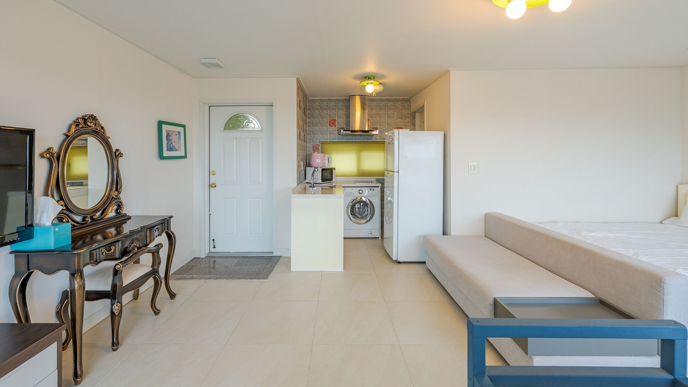 A compact room with a bed, a desk, a mirror, a washing machine, and a refrigerator.