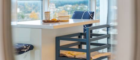 Una cocina moderna con encimera blanca, una silla azul y vistas al exterior a través de amplios ventanales.