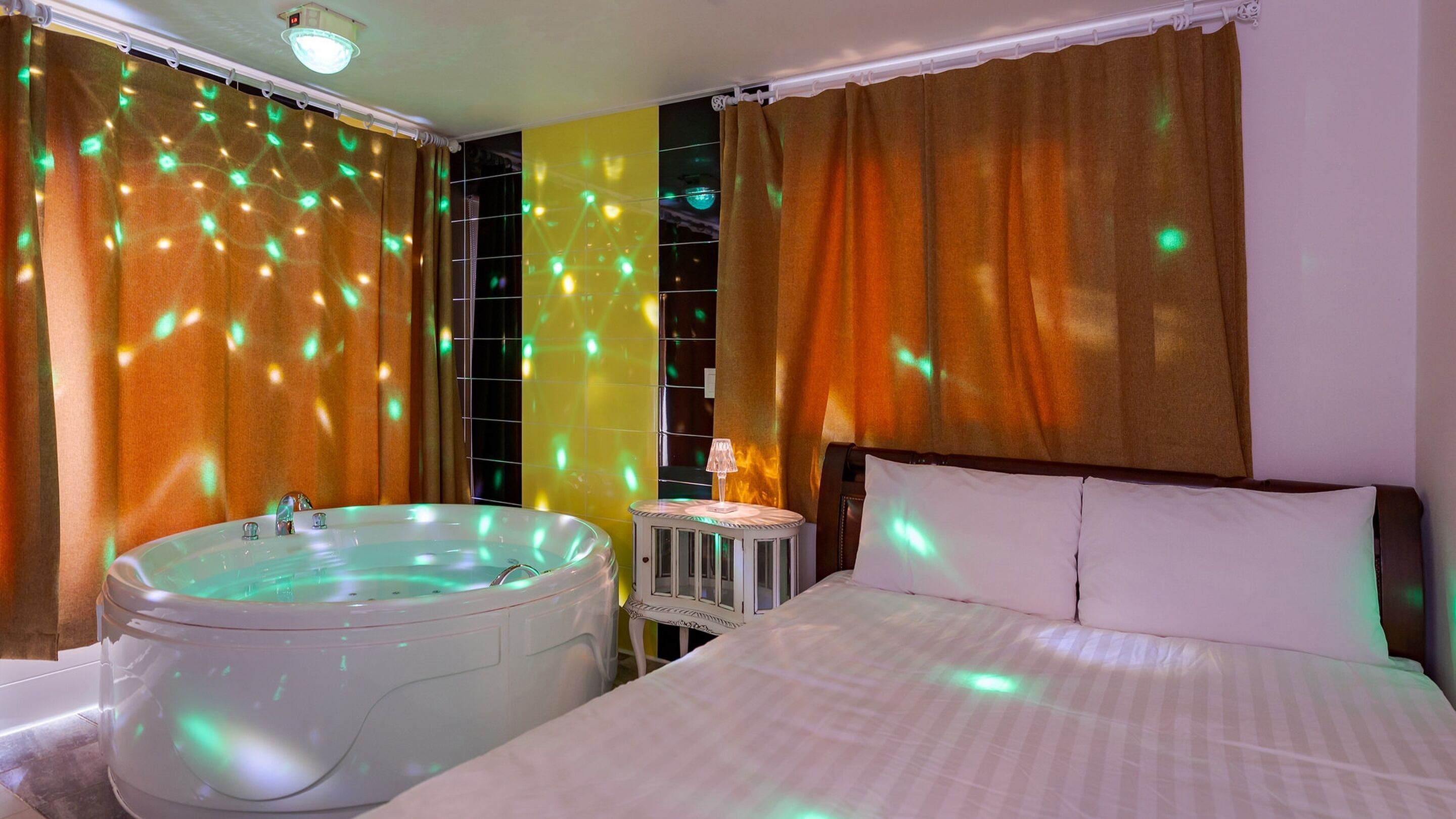A hotel room with a large bathtub, a bed with white linens, and curtains with light projections.
