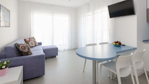 Un salon moderne comprenant un canapé, une table à manger avec des chaises, un téléviseur à écran plat fixé au mur et une fenêtre ornée de rideaux légers.