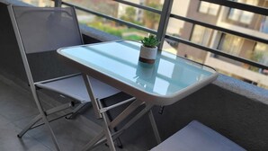 Uma varanda com mesa de vidro e duas cadeiras, um pequeno vaso com planta e vista para edifícios ao fundo.
