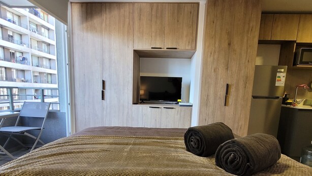 Quarto de hotel moderno com uma cama grande, televisão, cozinha compacta e varanda com vista para um edifício.