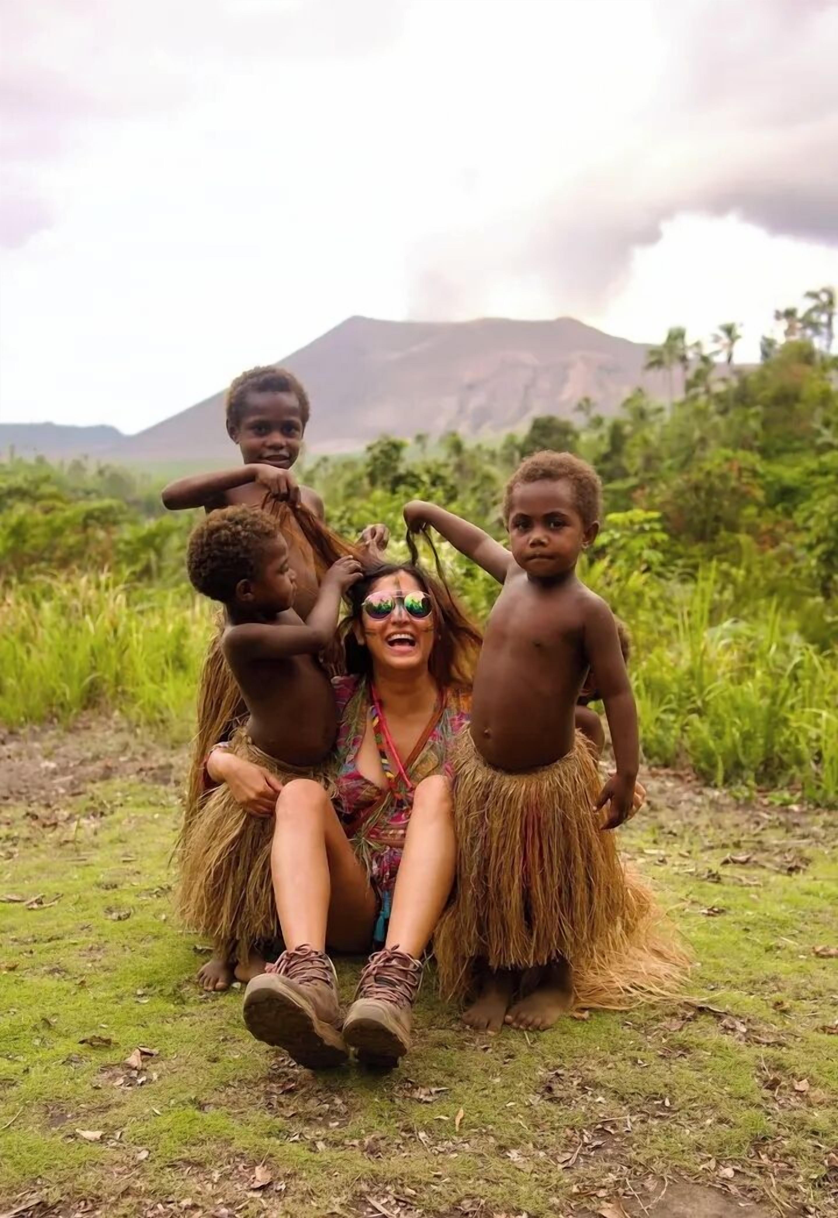 A person sitting on the ground with children around, in a natural outdoor setting with greenery and a mountain in the background.