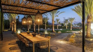 Un área de comedor al aire libre con una pérgola, sillas de mimbre, una mesa de madera y luces colgantes.
