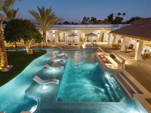 Une piscine avec plusieurs couloirs de nage, un espace détente avec des chaises et des parasols, et une terrasse bien éclairée.