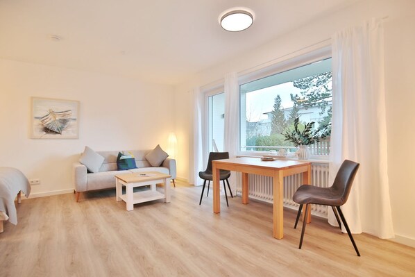 A modern living room with a sofa, a small table, and a dining table with chairs. There is a window with curtains, a radiator, and a painting on the wall.