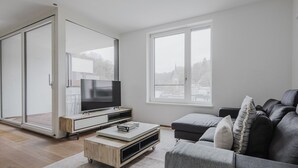 A modern living room with a sliding glass door leading to a balcony, a flat-screen TV on a wooden stand, a grey sofa, and a coffee table with books.