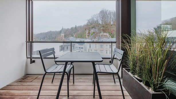A balcony with a table and chairs, a potted plant, and a view of buildings and trees.