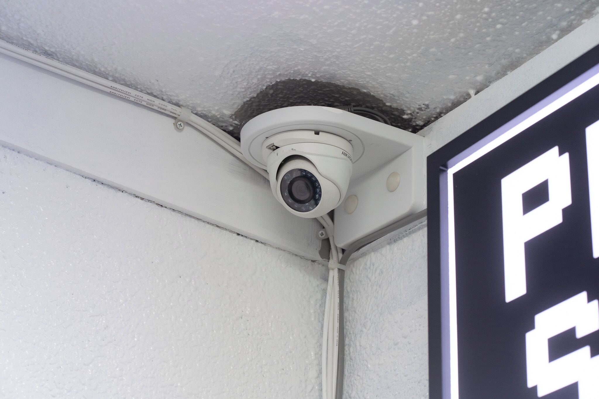 Una cámara de seguridad instalada en el techo, un letrero con las letras “P” y “C”, y una pared blanca con textura.