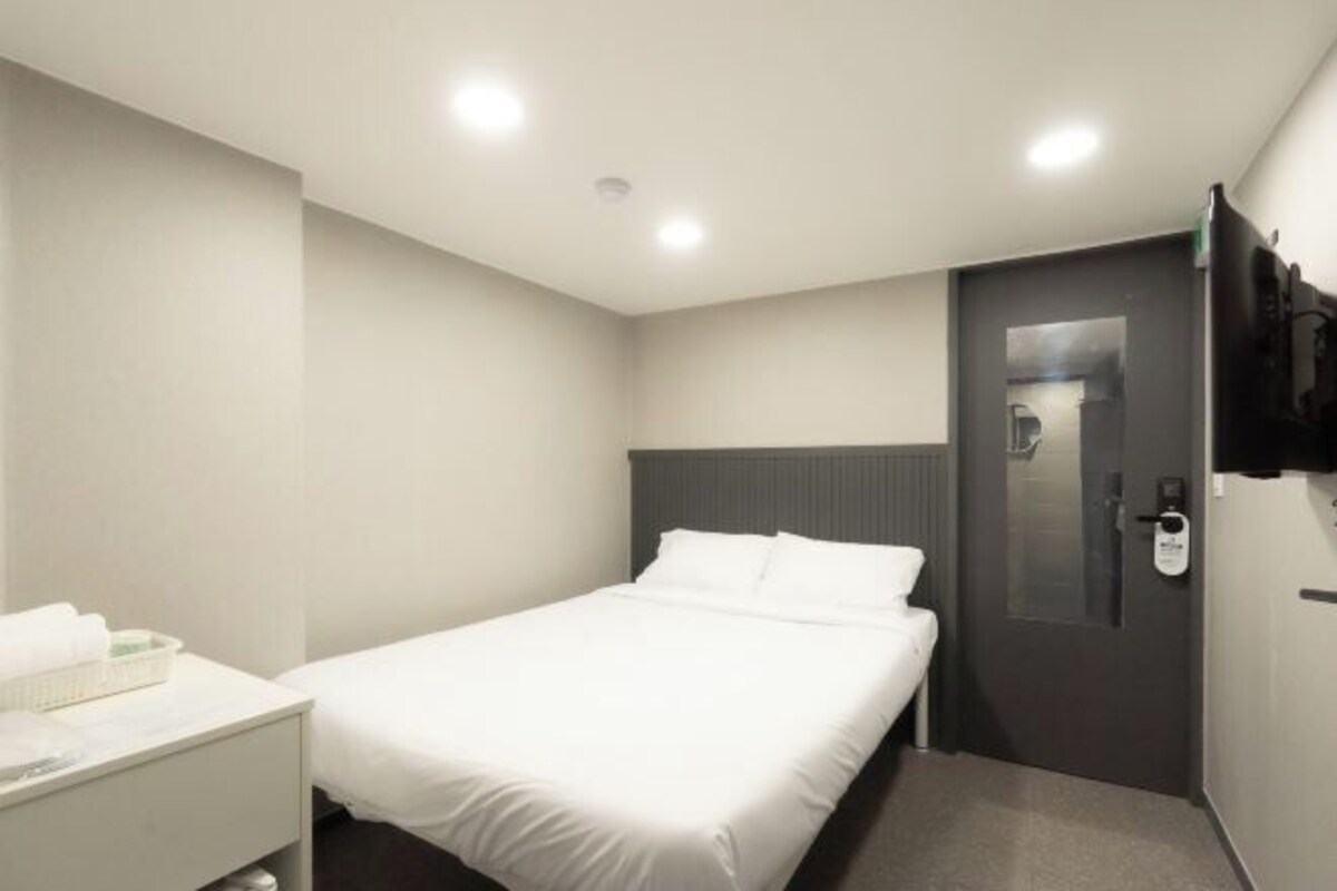 A modern hotel room with a single bed, a bedside table with a lamp, a flat-screen TV mounted on the wall, and a door with a handle.